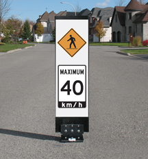 A vertical traffic sign installed in the centre of a residential street. The sign displays a yellow pedestrian crossing symbol above a white speed limit sign reading “Maximum 40 km/h.” Houses, trees, and a clear sky appear in the background.